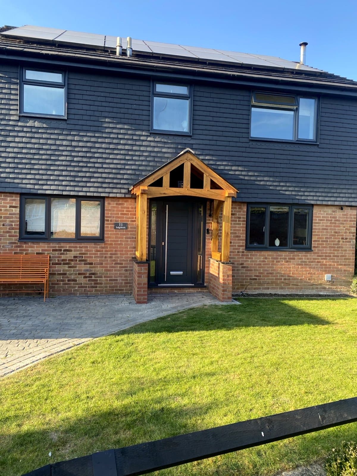 The Malvern oak porch on a contemporary home with dark cladding — brick plinths, curved braces and a tiled gable