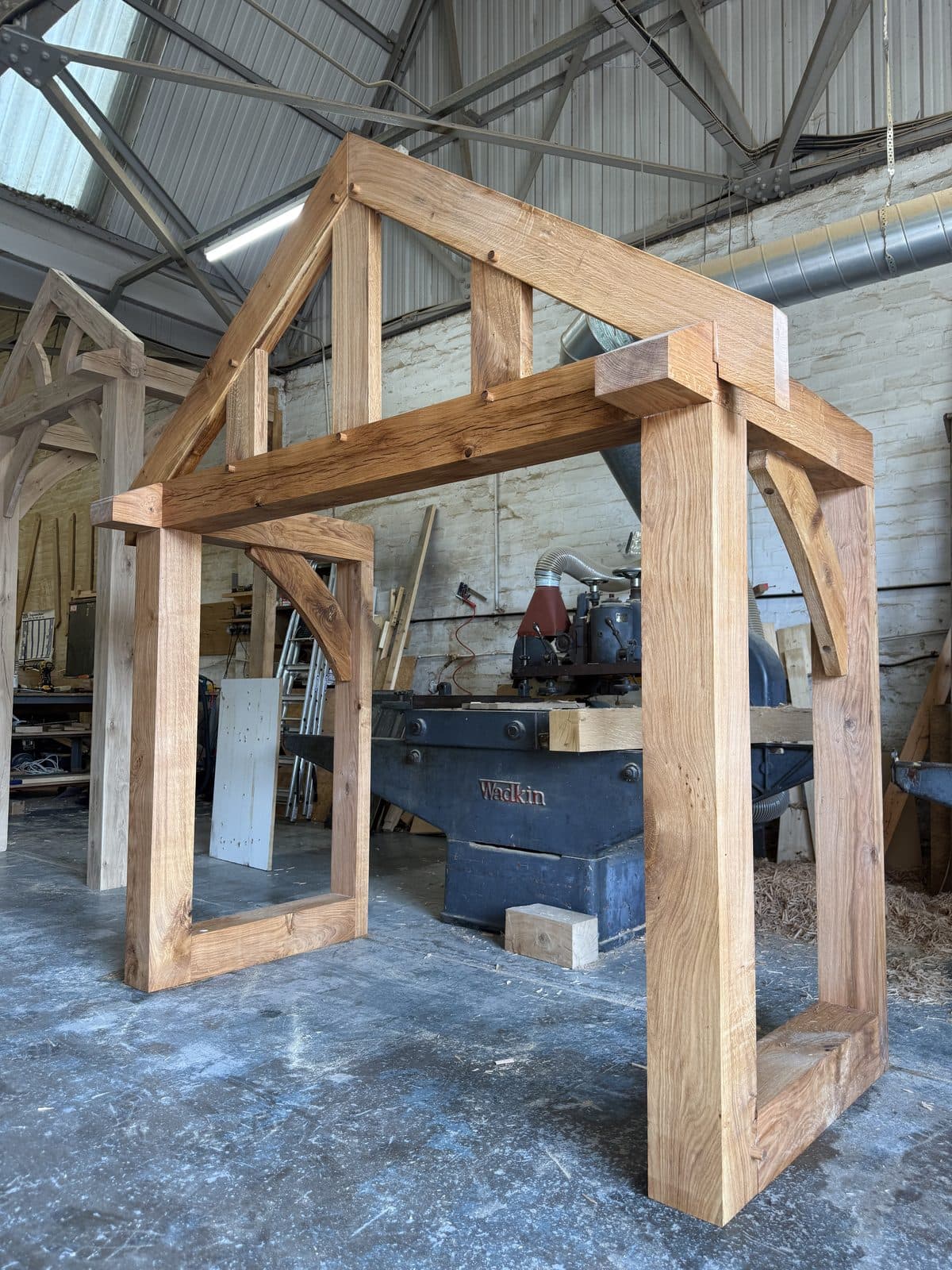 A freshly cut oak porch frame dry-fitted beside the Wadkin planer, curved braces pegged at the head of each post